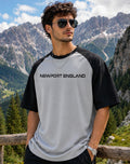 A man wearing a black and light gray raglan t-shirt with 'NEWPORT ENGLAND' printed across the chest, posing outdoors in front of a mountain backdrop.