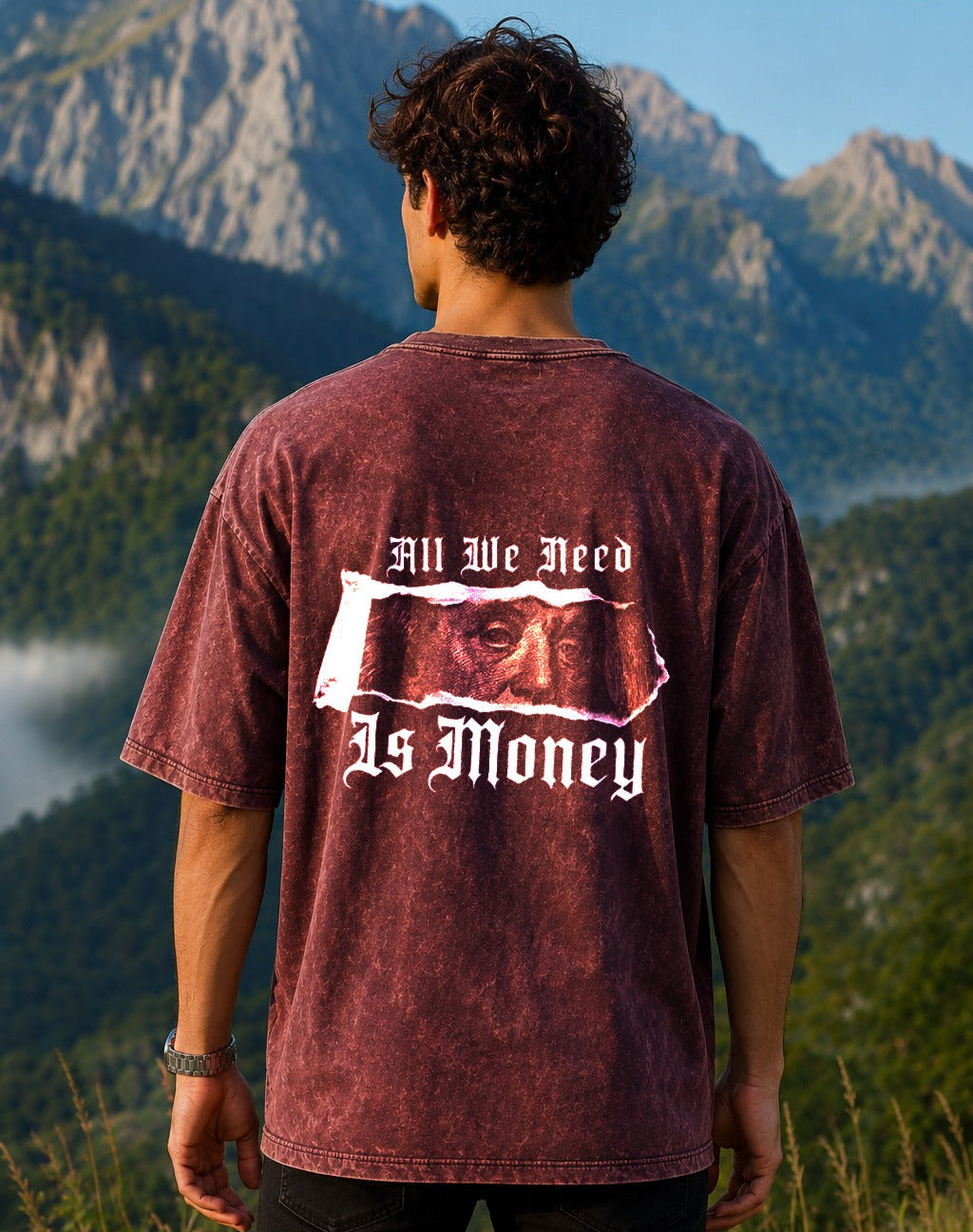 A man viewed from behind wearing a maroon acid-wash graphic t-shirt with a large white gothic text print reading 'All We Need Is Money' over a torn-paper graphic. The background shows a scenic mountain range.