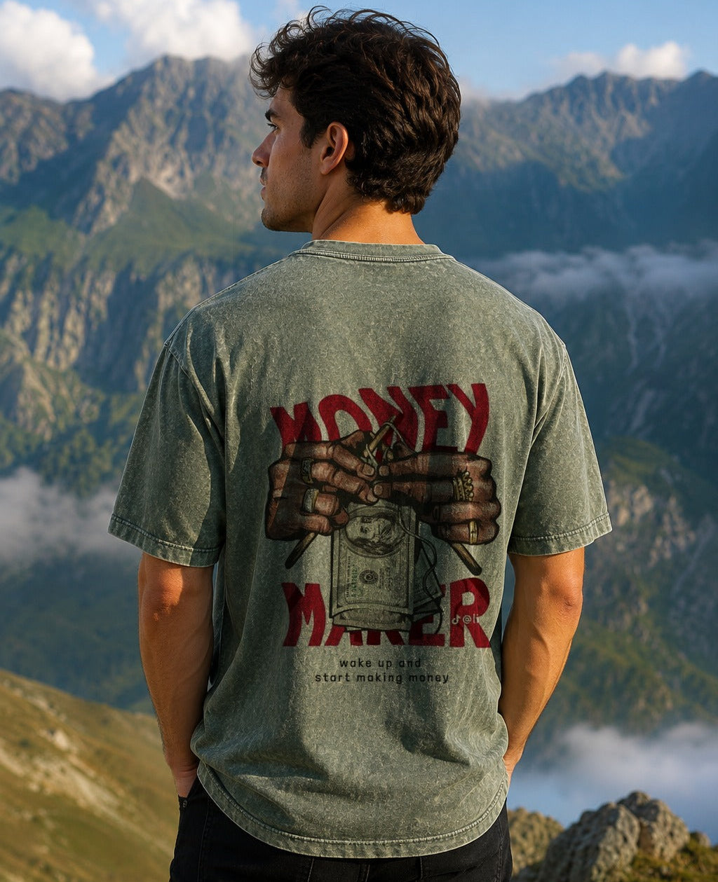 A man stands outdoors in front of mountains wearing a heathered sage green t-shirt with a large graphic on the back featuring the text 'MONEY MAKER' and an illustration of hands holding a stack of cash. The shirt has a visible 'True Money' brand logo printed below the graphic.