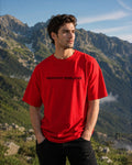 A man wearing a bright red t-shirt with 'NEWPORT ENGLAND' printed in black across the chest, standing outdoors against a backdrop of green and gray mountains under a blue sky.