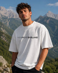 A man wearing a white crewneck t-shirt with 'NEWPORT ENGLAND' printed in black across the chest, set against a scenic mountain background.