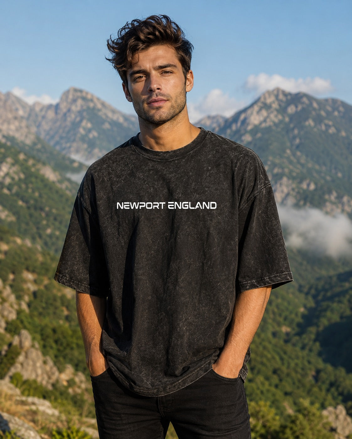 A man wearing a black acid-wash t-shirt with 'NEWPORT ENGLAND' printed in white across the chest, standing outdoors with a mountain landscape background.
