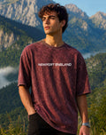 A man wearing a maroon acid-wash t-shirt with 'NEWPORT ENGLAND' printed in white across the chest, standing outdoors against a backdrop of green mountains and blue sky.
